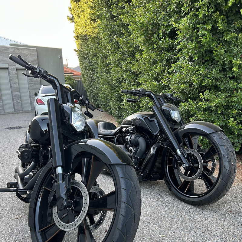 Two black custom motorcycles with large front wheels on a gravel driveway